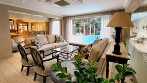 Amplia sala de estar con sofá, sillas, una mesa de centro y una lámpara. Se ve una piscina fuera.