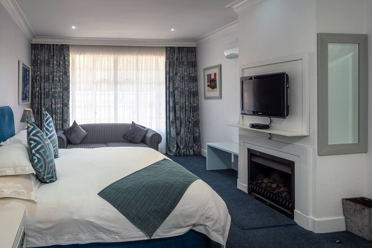 A modern hotel room with a large bed, a sofa, a flat-screen TV, a fireplace, and a large window with curtains.