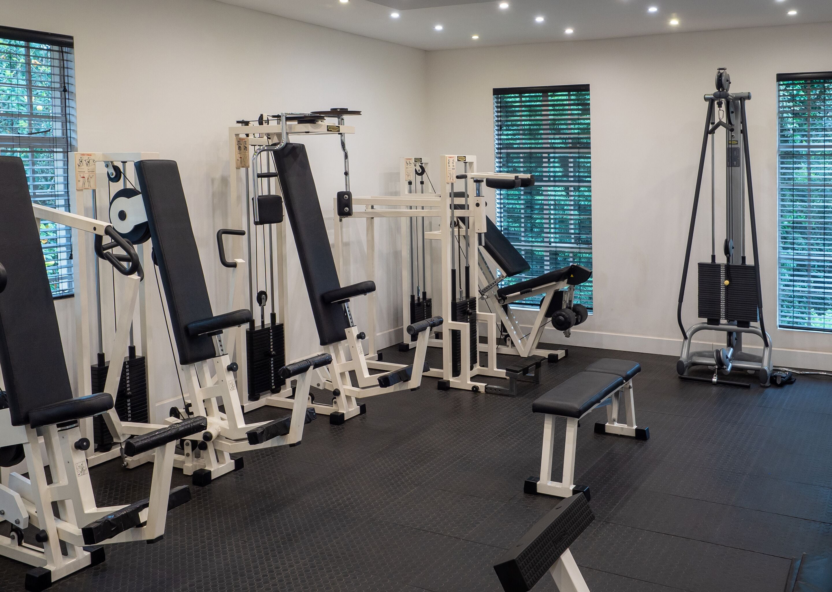 Ein Fitnessstudio mit verschiedenen Trainingsgeräten, einer Bank und einem Fenster mit Jalousien.