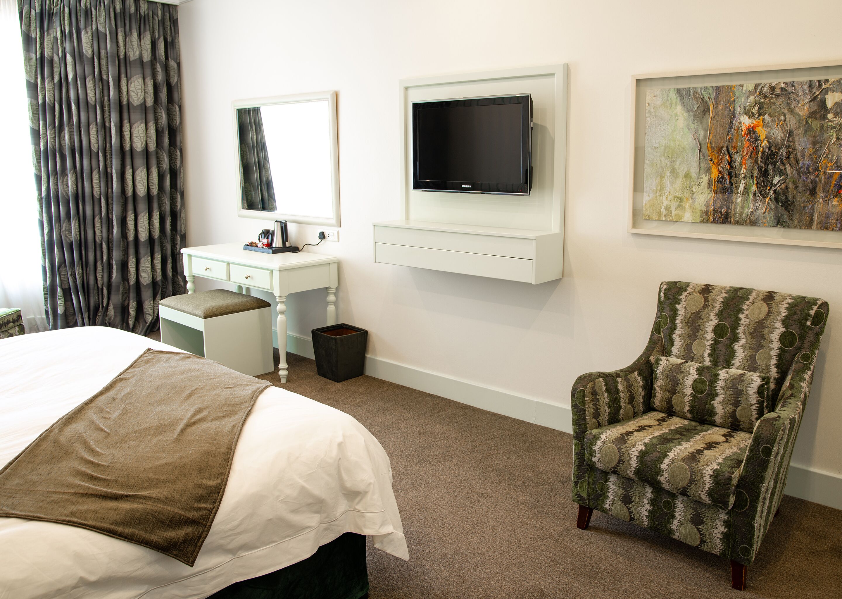 A hotel room with a bed, a desk, a chair, a TV, and a painting on the wall.