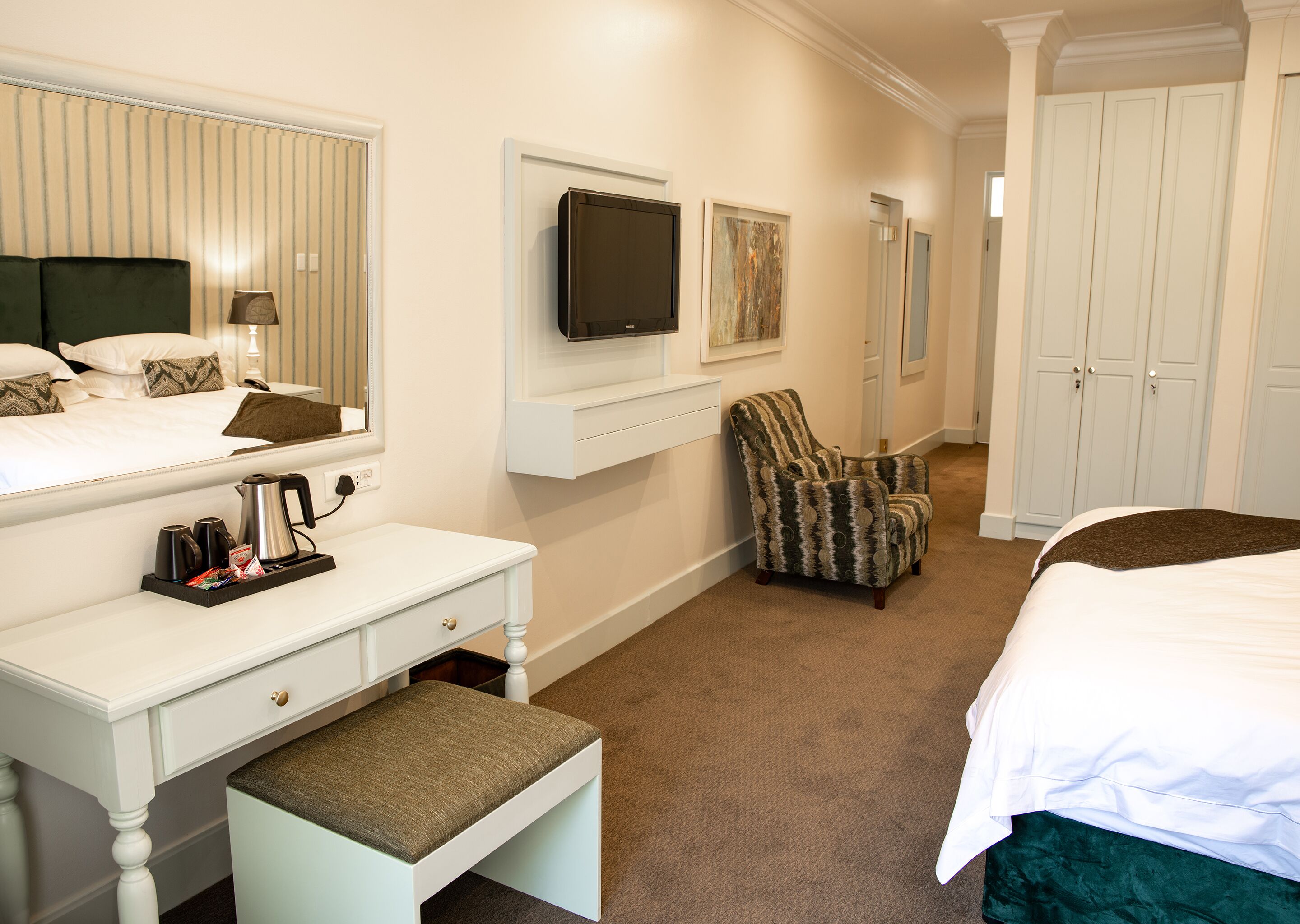 Quarto de hotel com cama, escrivaninha com cadeira, televisão e guarda-roupa.