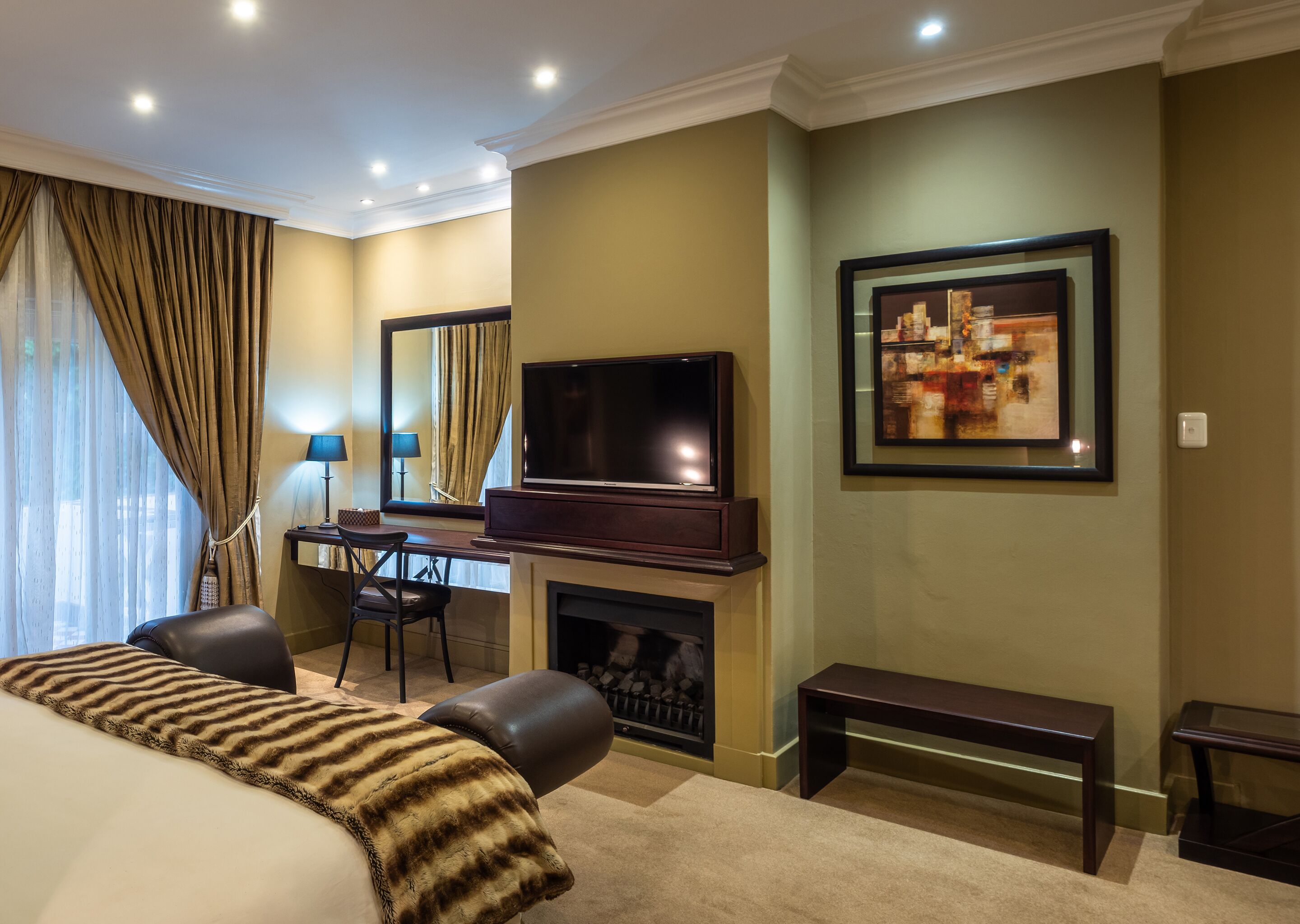 A hotel room with a large bed, a desk with a chair, a television, a fireplace, and a framed picture on the wall.