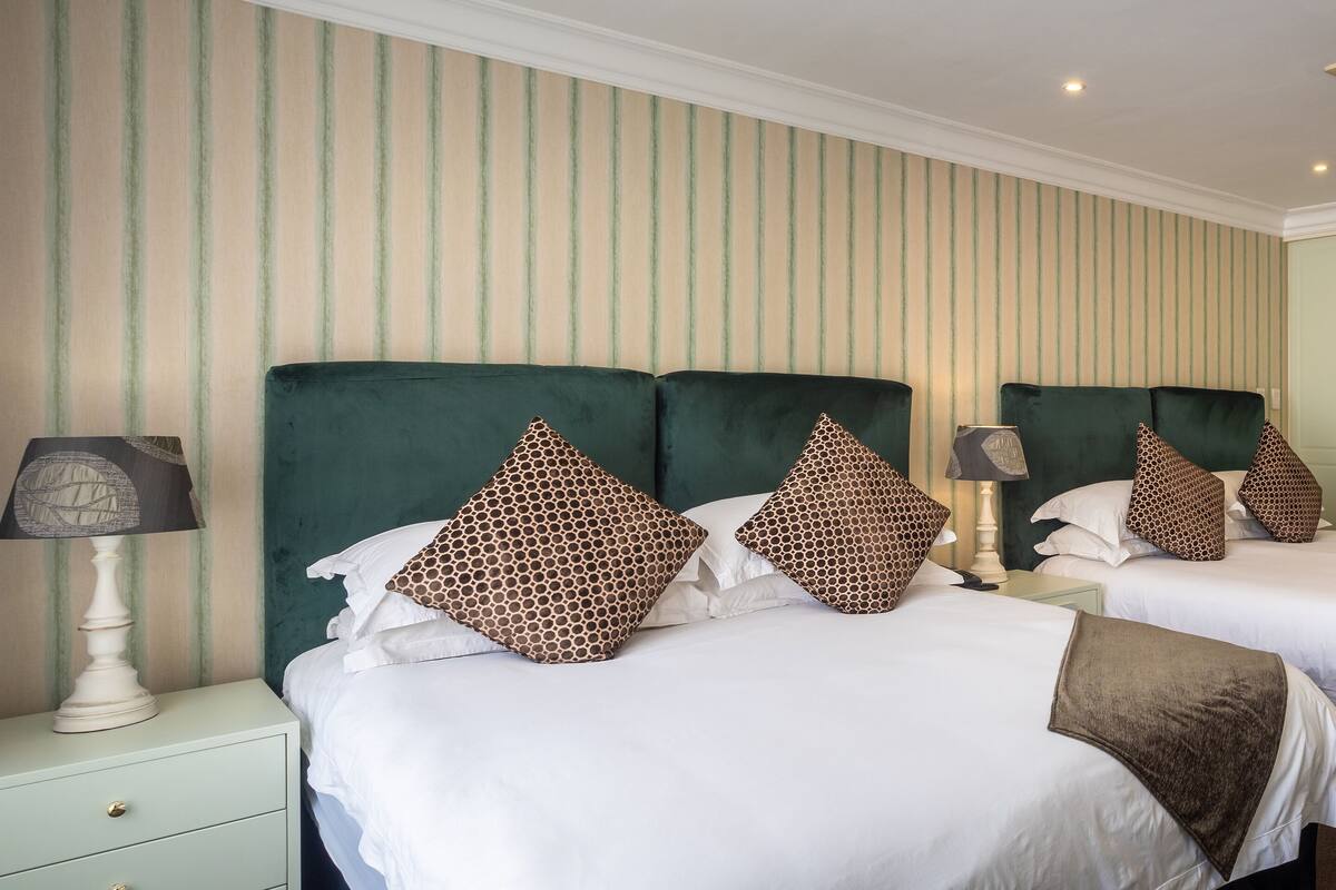 A hotel room with two beds, each with green upholstered headboards and white bedding. There are patterned pillows and a lamp on each nightstand.