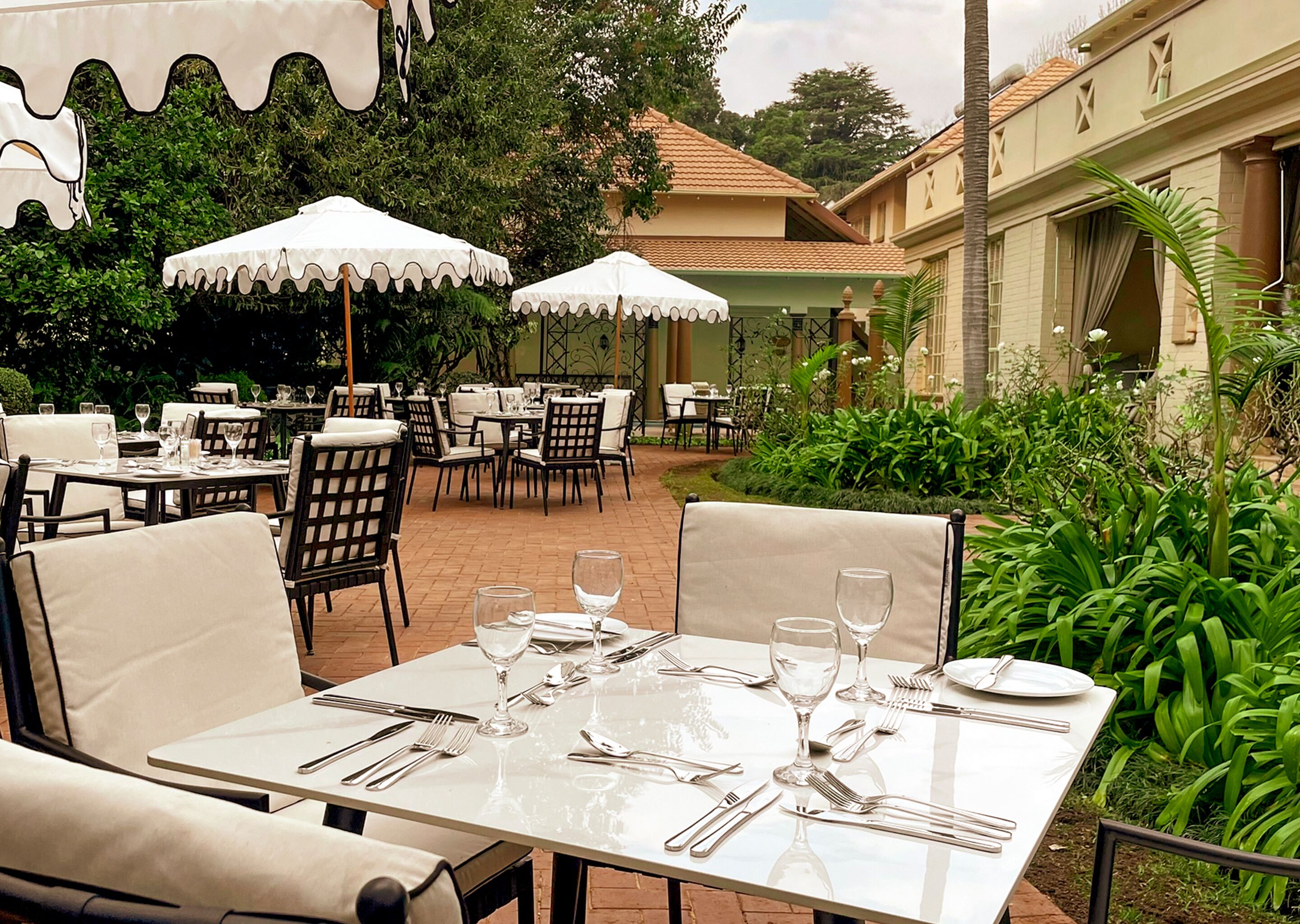 An outdoor dining area with tables and chairs, surrounded by greenery and a building with a tiled roof.