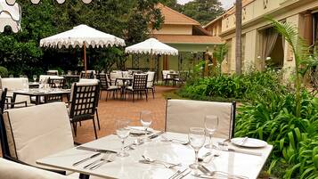 An outdoor dining area with tables and chairs, surrounded by greenery and a building with a tiled roof.