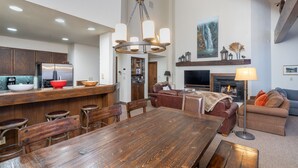 A spacious living room with a fireplace, a dining area with a wooden table, and a kitchen with wooden cabinets and a refrigerator. - Kayenta 7-Curate Telluride, Perfect SkI In/Out, Lift 4, Hot Tub, Fireplace (Mountain Village)