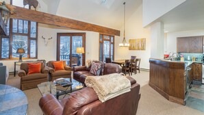 A spacious living area with leather sofas, a glass coffee table, and a fireplace. There is a large window with a view of the outdoors, and a kitchen area with wooden cabinets and a bar. - Kayenta 7-Curate Telluride, Perfect SkI In/Out, Lift 4, Hot Tub, Fireplace (Mountain Village)