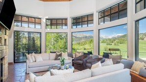 A spacious living room with a large sofa, armchairs, and a coffee table. Large windows offer views of a mountainous landscape.