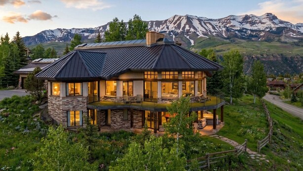 A multi-story house with a stone facade, large windows, and a balcony overlooking a mountainous landscape.