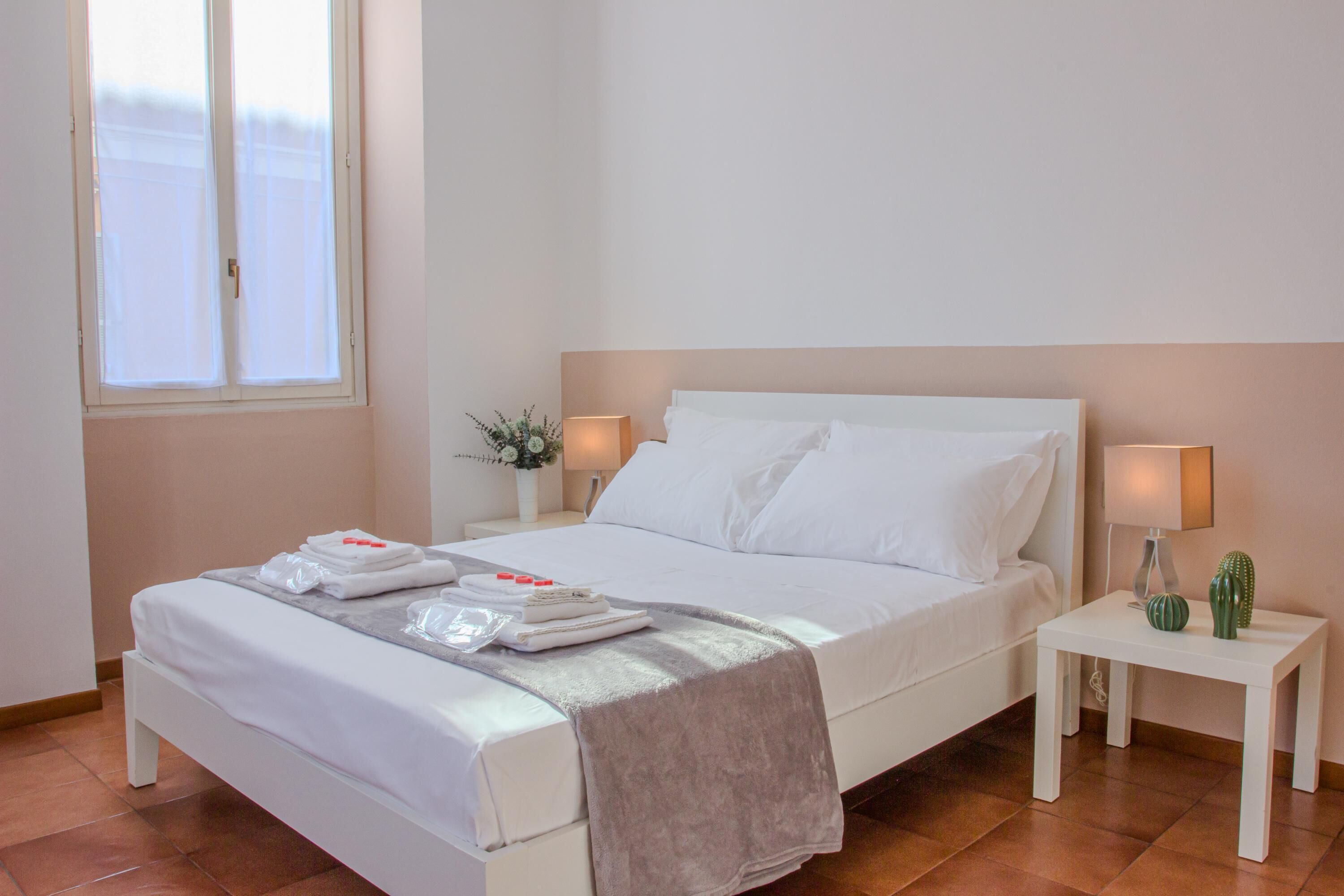 Een net opgemaakt bed met witte lakens, een nachtkastje met een lamp en een kleine cactus.