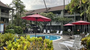 Un espace piscine doté de chaises longues et de parasols, entouré d’une végétation luxuriante et bordé d’un immeuble à plusieurs étages avec des balcons.