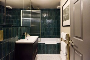 A modern bathroom with a dark green tiled wall, a white sink, a bathtub, and a framed artwork above the mirror.