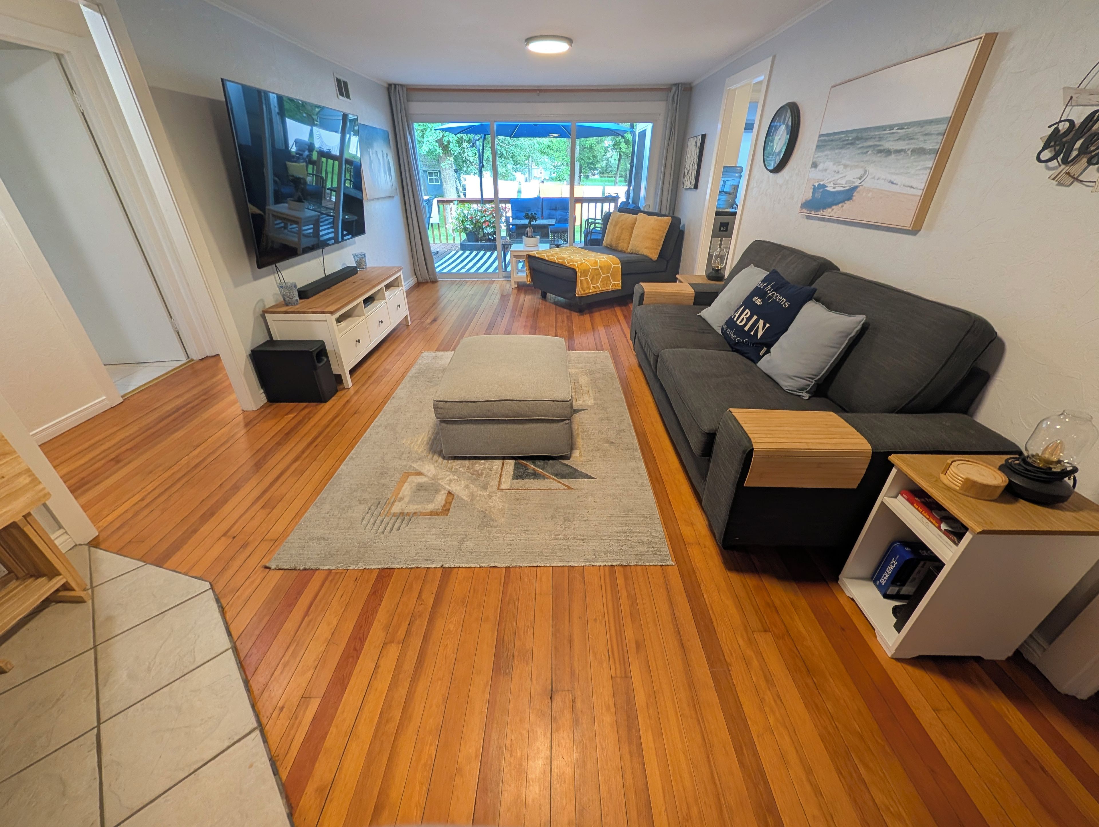 A modern living room with a sofa, ottoman, and a TV. There is a view of a balcony with outdoor furniture.
