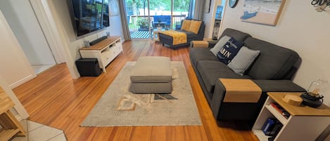 A modern living room with a sofa, ottoman, and a TV. There is a view of a balcony with outdoor furniture.