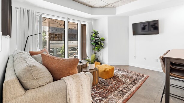 A modern living room with a sofa, a small side table with a plant, a yellow ottoman, and a patterned rug. - Le Downtown (rooftop pool and gym) (Québec)