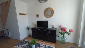A modern living room with a black entertainment unit, a television, a patterned rug, a potted plant, and a vase with flowers. - - L'olivier - (Dozulé)