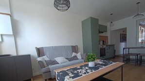 A modern living room with a grey sofa, a coffee table with a floral table runner, a grey cabinet, and a kitchen area in the background. - - L'olivier - (Dozulé)