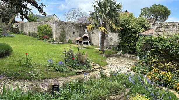 Un cortile con pareti in pietra, un prato verdeggiante, un piccolo giardino con diverse piante e fiori, e un forno in pietra tradizionale.