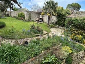 Ein Innenhof mit Steinmauern, einer grünen Rasenfläche, einem kleinen Garten mit verschiedenen Pflanzen und Blumen sowie einem traditionellen Steinofen.