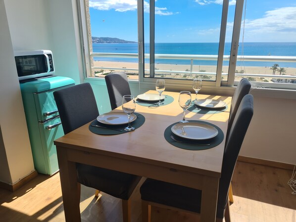 Una mesa de comedor con platos y copas de vino, un refrigerador color turquesa y vista a la playa.