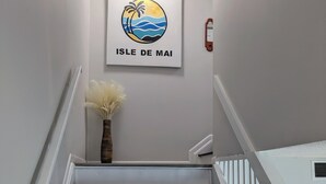 Una escalera con escalones negros y barandas blancas, un cuadro enmarcado con una playa, y un jarrón con flores blancas.