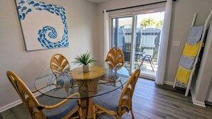 Un comedor con una mesa de vidrio rodeada de sillas de mimbre, una planta en maceta y un cuadro abstracto azul en la pared.