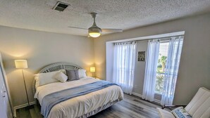 Un dormitorio con cama, ventilador de techo, mesita de noche, una silla y una ventana con cortinas.