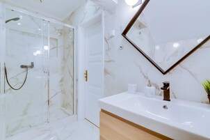A modern bathroom with a glass shower enclosure, a marble-tiled floor, a white sink with a wooden cabinet, and a large mirror with a gold frame.