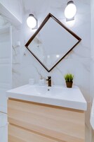 Un baño con un lavamanos de mármol blanco, un mueble de tocador de madera, un espejo con marco marrón y dos luces empotradas.