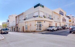 Un edificio de varios pisos con acceso en planta baja, un estacionamiento con coches y un cielo despejado.