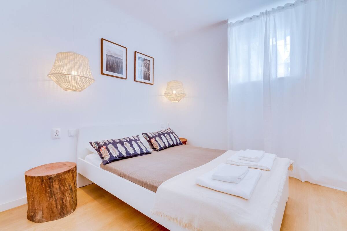 Quarto minimalista com cama, duas luminárias fixadas na parede e janela com cortinas leves.