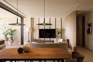 A modern living room with a wooden dining table, a sofa, a flat-screen TV mounted on the wall, and pendant lighting.