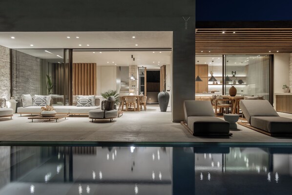 A modern living area with a pool, featuring a sofa set, dining area, and a kitchen visible through glass doors.
