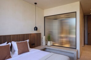 A modern hotel room with a bed, a bedside table with a plant, a large window with blinds, and a hanging light fixture.