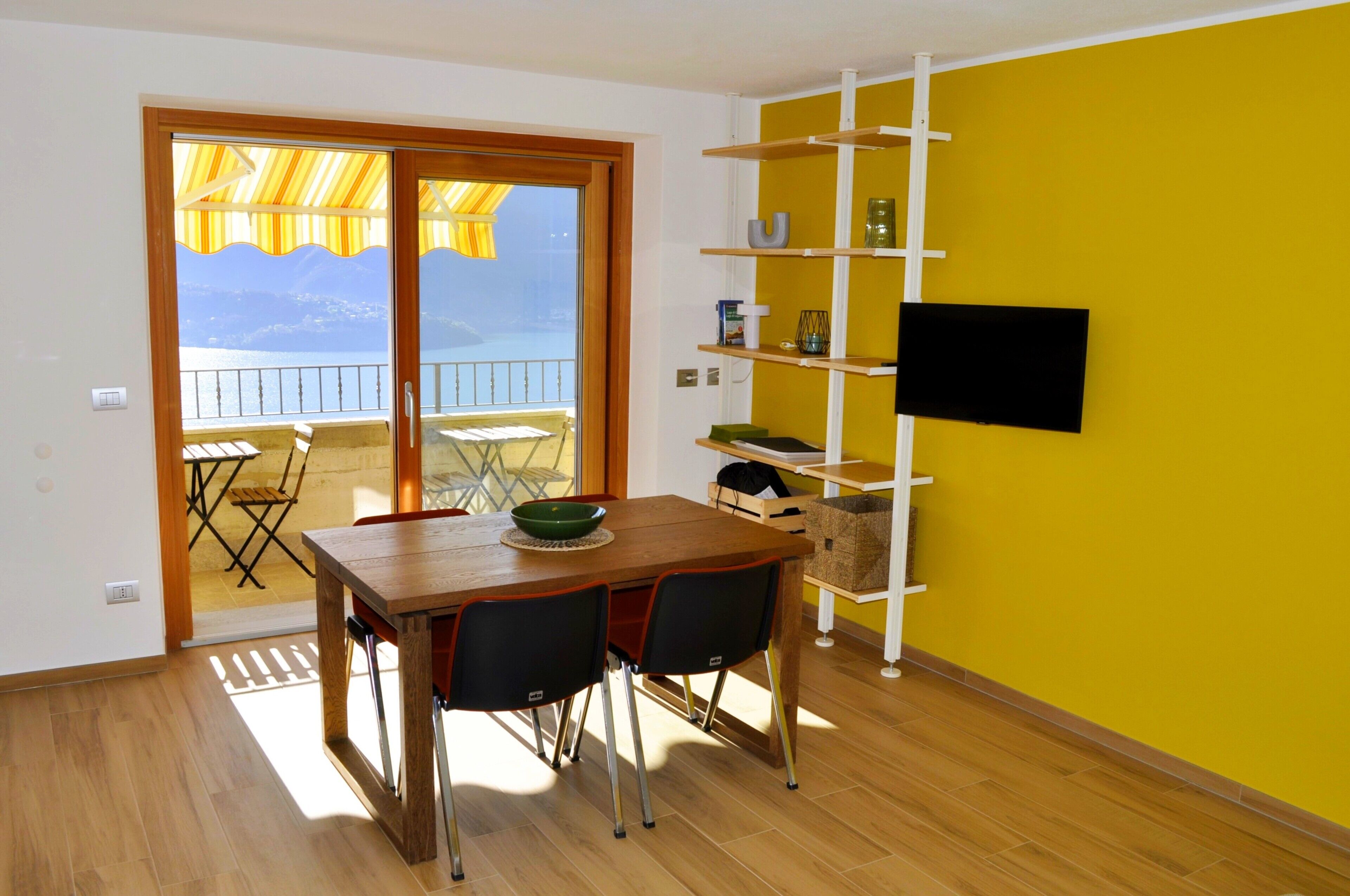 A dining area with a wooden table and chairs, a television mounted on a yellow wall, and a balcony with a view of the sea.