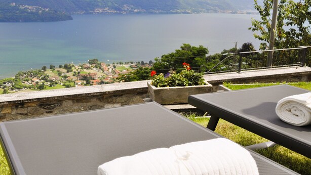 Una terraza con vistas a un lago y montañas, una silla de descanso con una toalla y una jardinera con flores.