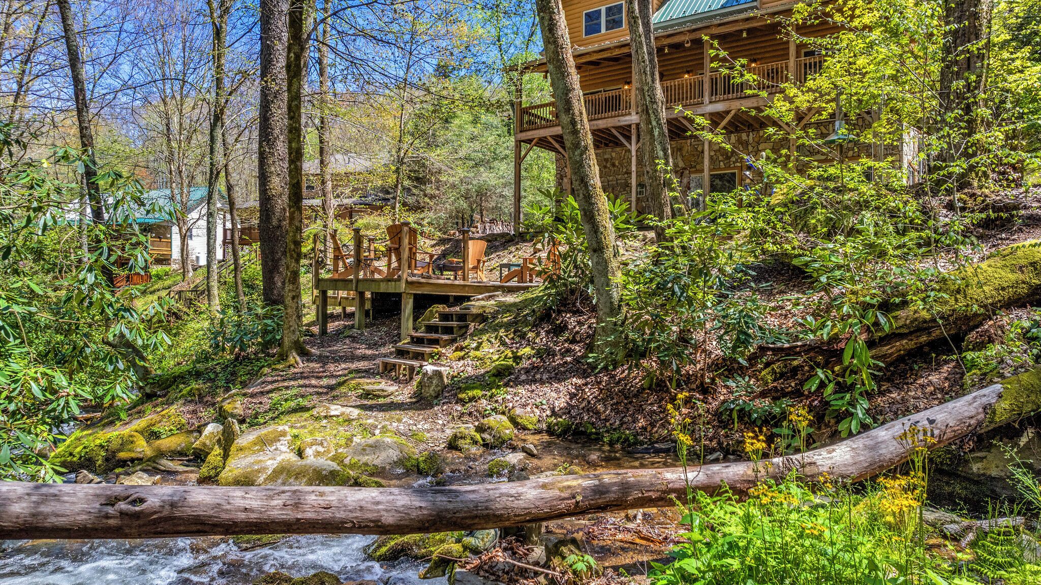 A wooden cabin with a stone foundation, surrounded by a forested area with a small stream.