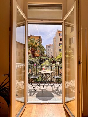 Een open deur die leidt naar een balkon met een tafel en stoelen, omringd door groen en gebouwen op de achtergrond.