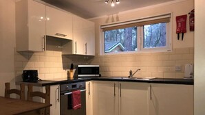 A modern kitchen with white cabinets, a microwave, a stove, and a window with a view of a house. - Cosy Cabin (Finchampstead)