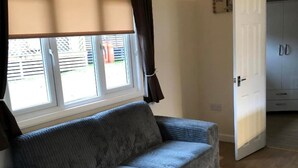 A room with a grey sofa, a window with blinds, a wooden floor, and a door leading to another room. - Cosy Cabin (Finchampstead)