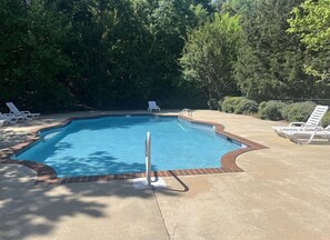 A rectangular swimming pool with a brick border, surrounded by a concrete deck. There are lounge chairs along the pool's edge.