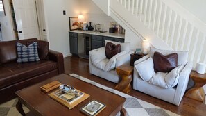 Un salon avec un canapé en cuir marron, deux fauteuils blancs avec des coussins marron, une table basse en bois sur laquelle sont posés des livres et un plateau, et un escalier menant à un espace cuisine.