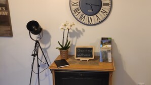 Une pièce avec une table en bois sur laquelle se trouvent une lampe noire, une plante en pot, une pancarte indiquant « Bienvenue », un téléphone intelligent et un livre. Une grande horloge murale avec des chiffres romains est accrochée au-dessus de la table.
