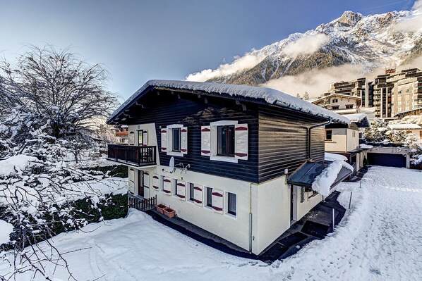 A chalet-style building with a balcony, surrounded by snow-covered trees and mountains in the background.