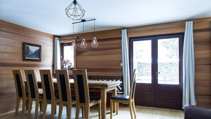 Et rum med træpaneler, et spisebord med stole, en hængende lampe og store vinduer med gardiner.