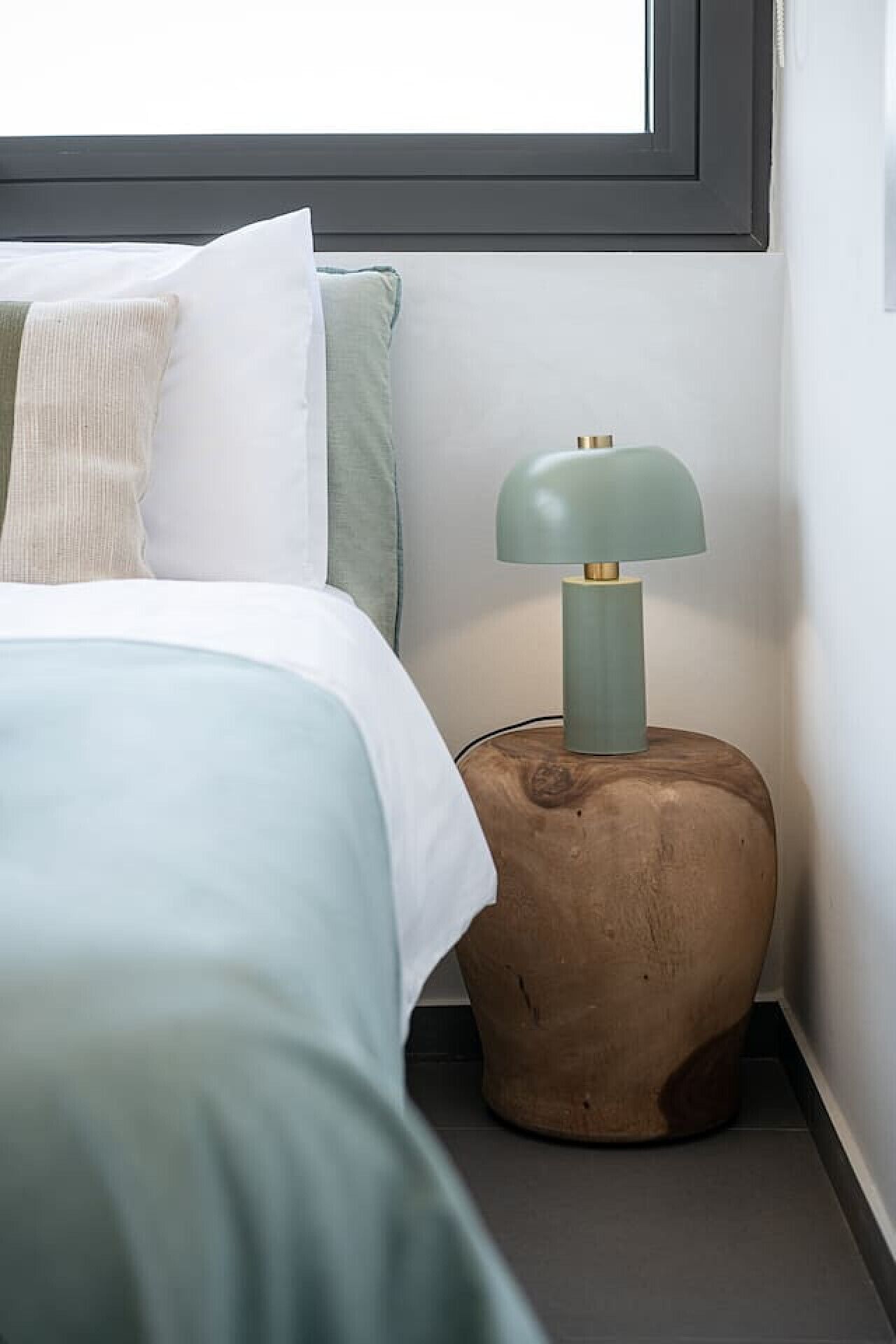 A neatly made bed with a green and white duvet, a wooden nightstand with a green lamp, and a window above the bed.