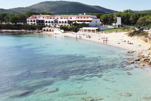 A coastal hotel with a large building, a sandy beach, and clear turquoise waters.