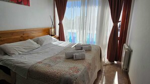 A hotel room with a wooden headboard, a bed with white linens, a floral-patterned bedspread, and two towels on the bed. - COMPLEJO RAICES PATAGONICAS DEPARTAMENTO 2 HABITACIONES CON BALCON (Dina Huapi)