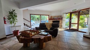 A spacious living room with a fireplace, a large window, and a chessboard on a wooden coffee table. - COMPLEJO RAICES PATAGONICAS DEPARTAMENTO 2 HABITACIONES CON BALCON (Dina Huapi)