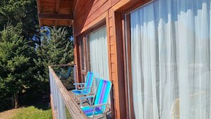 A wooden cabin with a balcony, two striped chairs, and a large window with sheer curtains. - COMPLEJO RAICES PATAGONICAS DEPARTAMENTO 2 HABITACIONES CON BALCON (Dina Huapi)